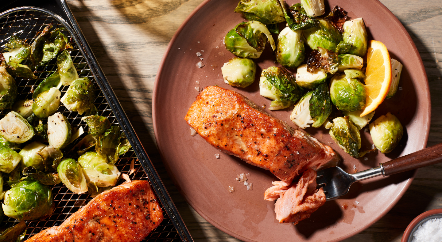 Air fryer salmon and Brussel sprouts on a plate next to an air fry basket with salmon and Brussel sprouts. Air fryer salmon and Brussel sprouts on a plate next to an air fry basket with salmon and Brussel sprouts.