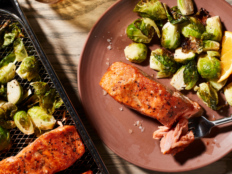 Air fryer salmon and Brussel sprouts on a plate next to an air fry basket with salmon and Brussel sprouts. Air fryer salmon and Brussel sprouts on a plate next to an air fry basket with salmon and Brussel sprouts.