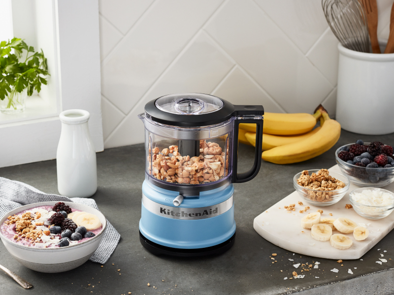 Yogurt and fruit next to a blue KitchenAid® food processor with nuts inside Yogurt and fruit next to a blue KitchenAid® food processor with nuts inside