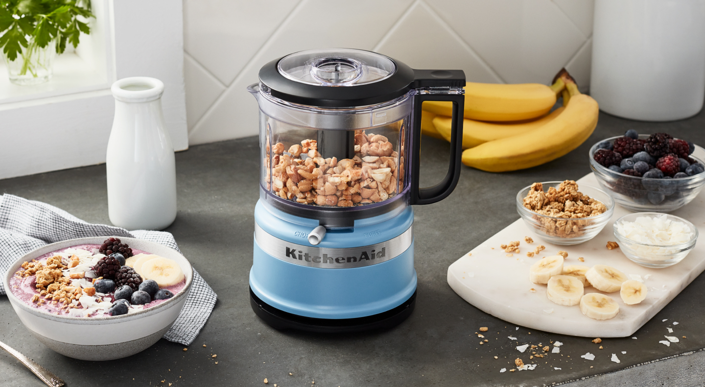 Yogurt and fruit next to a blue KitchenAid® food processor with nuts inside Yogurt and fruit next to a blue KitchenAid® food processor with nuts inside