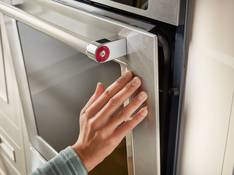A hand touching a stainless steel oven door with a glass window in it A hand touching a stainless steel oven door with a glass window in it