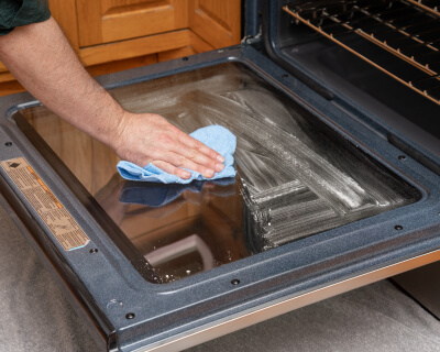 The inside of oven door glass with a hand wiping it down with a towel in the foreground