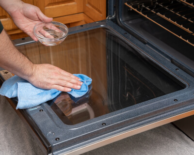 The inside of oven door glass with a hand wiping it down with a cloth treated with a vinegar or lemon mixture in the foreground