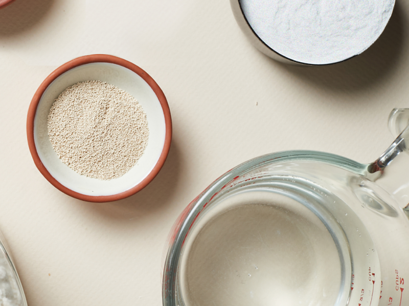 Dry ingredients like yeast and flour in small bowls next to a measuring cup with water Dry ingredients like yeast and flour in small bowls next to a measuring cup with water