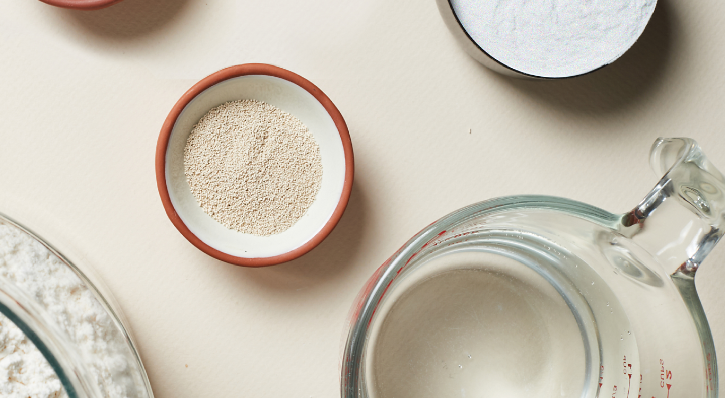 Dry ingredients like yeast and flour in small bowls next to a measuring cup with water Dry ingredients like yeast and flour in small bowls next to a measuring cup with water