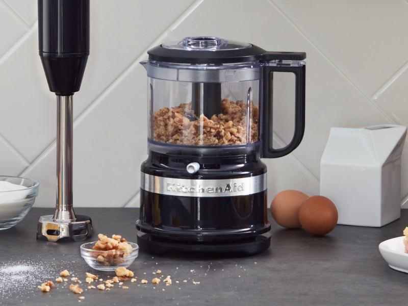 Black KitchenAid® food processor filled with walnuts sitting next to an upright immersion blender on a counter  Black KitchenAid® food processor filled with walnuts sitting next to an upright immersion blender on a counter