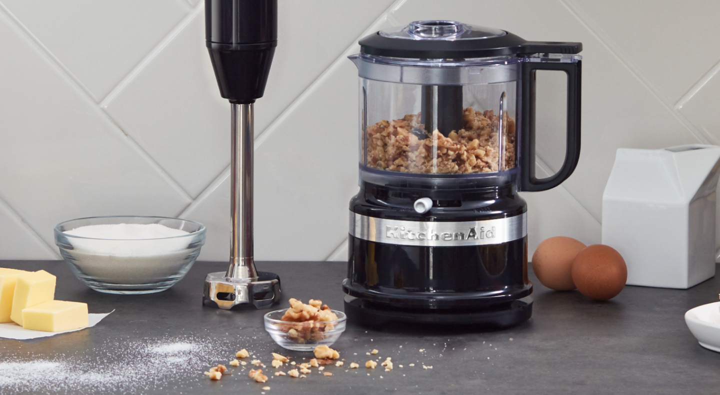 Black KitchenAid® food processor filled with walnuts sitting next to an upright immersion blender on a counter  Black KitchenAid® food processor filled with walnuts sitting next to an upright immersion blender on a counter