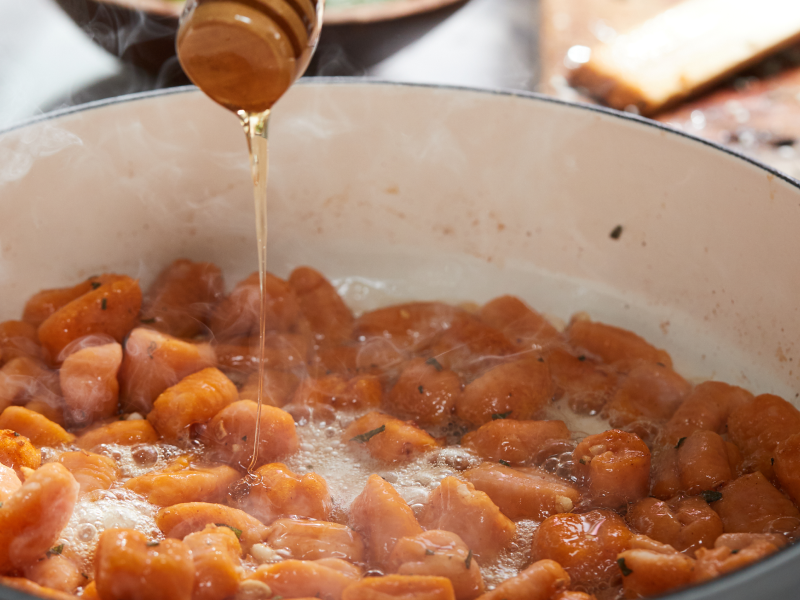 A wooden honey stick drizzling honey over sweet potato gnocchi sauteing on the stovetop.