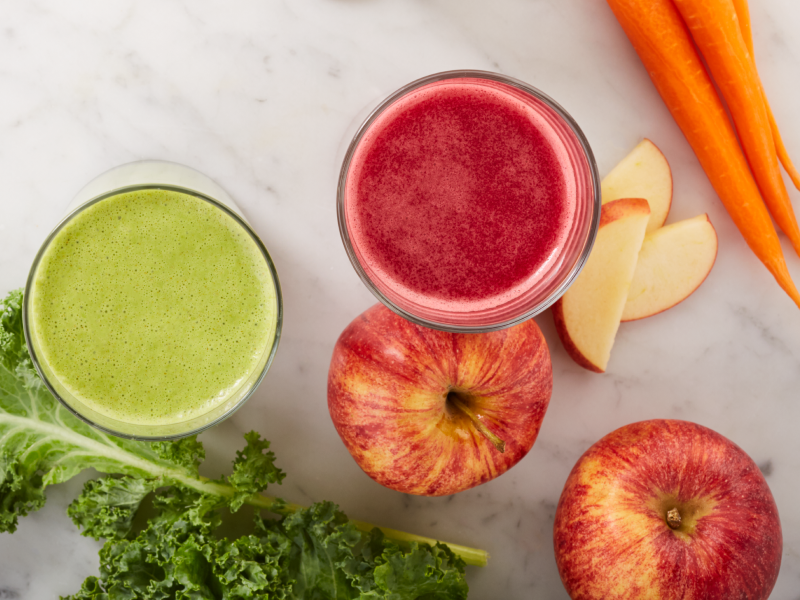 Green and red juices next to carrots, apples and lettuce on counter Green and red juices next to carrots, apples and lettuce on counter