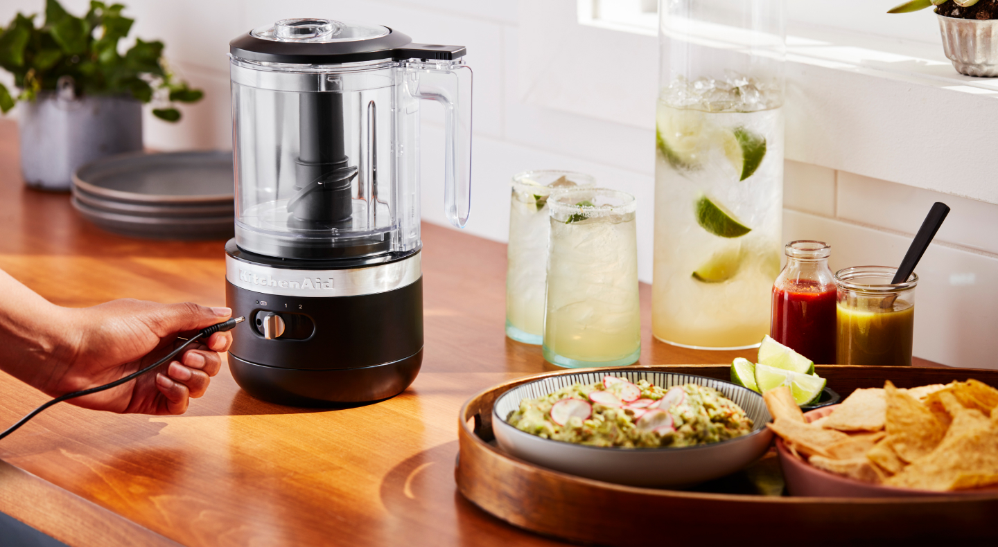 A cordless KitchenAid® food chopper next to a platter of guacamole and tortilla chips and a pitcher and glasses of margaritas.  A cordless KitchenAid® food chopper next to a platter of guacamole and tortilla chips and a pitcher and glasses of margaritas. A cordless KitchenAid® food chopper next to a platter of guacamole and tortilla chips and a pitcher and glasses of margaritas.