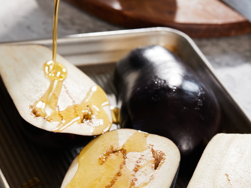 Olive oil drizzling on halved eggplants laying on baking sheet