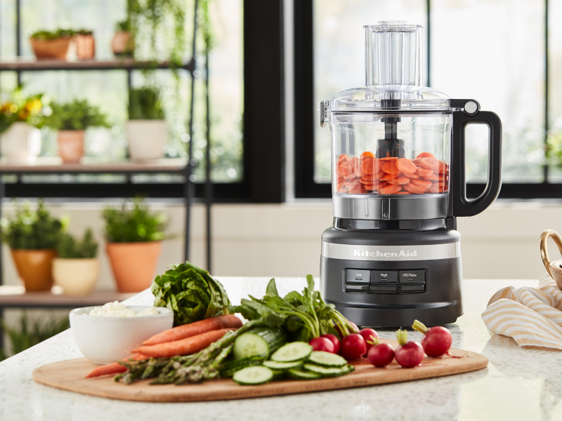 Cucumbers, carrots and radishes on cutting board next to food processor filled with sliced carrots Cucumbers, carrots and radishes on cutting board next to food processor filled with sliced carrots