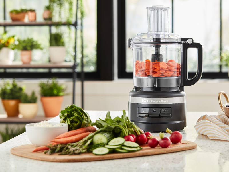 A KitchenAid® food processor mixing vegetables in a bright, modern kitchen. A KitchenAid® food processor mixing vegetables in a bright, modern kitchen.