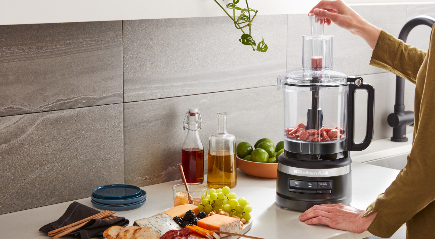 Person dropping food into the feed tub of a black KitchenAid® food processor Person dropping food into the feed tub of a black KitchenAid® food processor