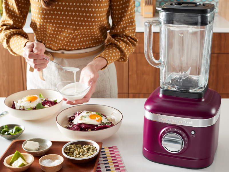 Person topping a harvest bowl with sauce next to a KitchenAid® blender Person topping a harvest bowl with sauce next to a KitchenAid® blender
