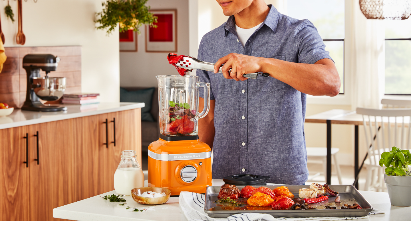 Person adding roasted vegetables into an orange KitchenAid® blender Person adding roasted vegetables into an orange KitchenAid® blender