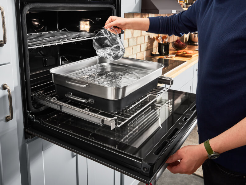 A person putting water in steaming cookware in the oven