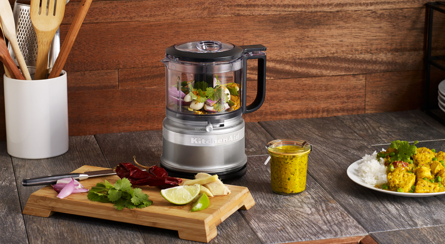 Various prepped ingredients on wood block cutting board next to full silver food chopper