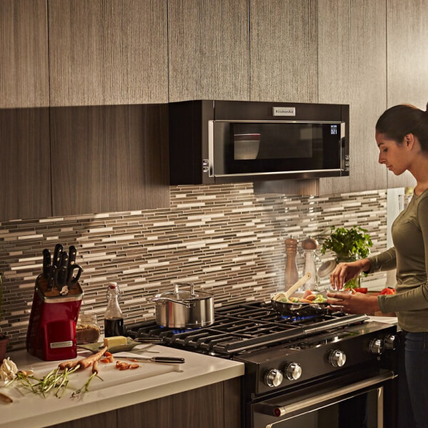 Person preparing stovetop recipe while food heats in a KitchenAid® low profile microwave above