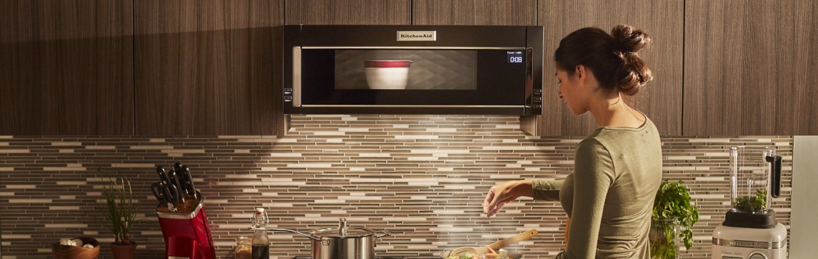 Person preparing a recipe on a gas stovetop below a KitchenAid® low profile microwave