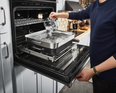 A person adding water to a broiler tray in an open oven