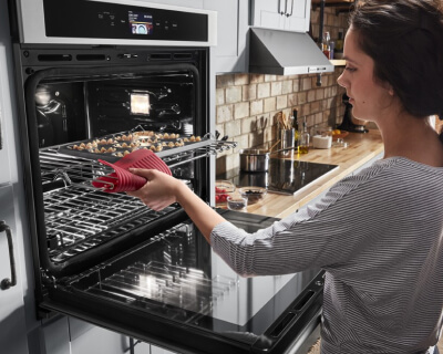 A woman pulling a baking tray out of a wall oven with a read potholder