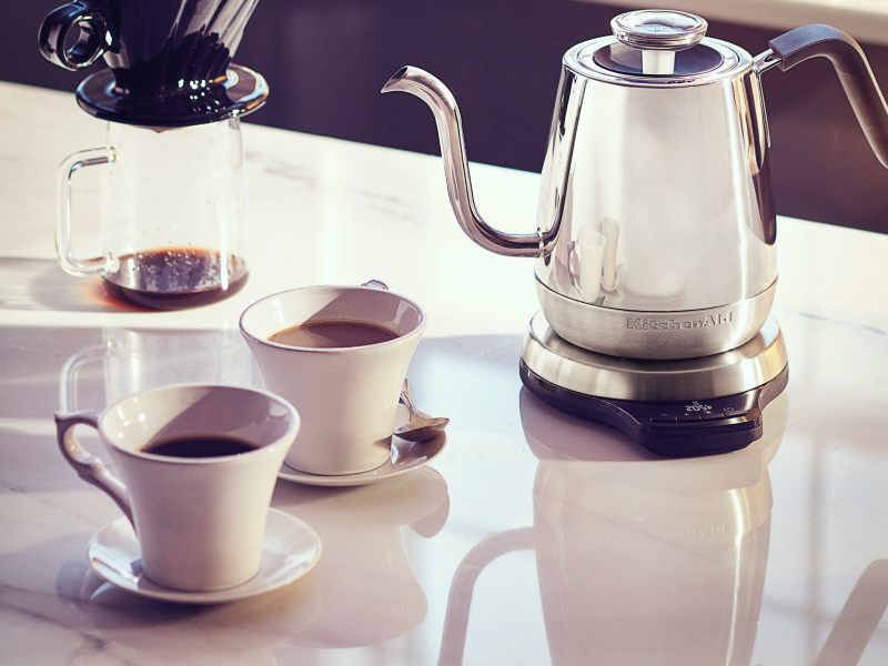 Silver KitchenAid® gooseneck electric kettle on white countertop with pour over coffee container and two cups of coffee