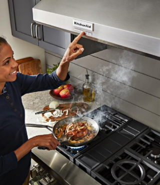 A person cooking on a KitchenAid® range, turning on the fan of a range hood