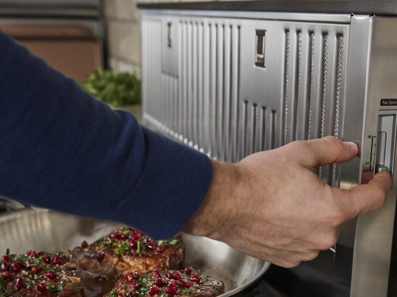 Man turning on a downdraft vent while meat cooks on the stove Man turning on a downdraft vent while meat cooks on the stove