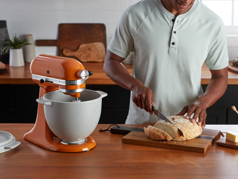 Person slicing crispy bread on cutting board next to orange KitchenAid® stand mixer