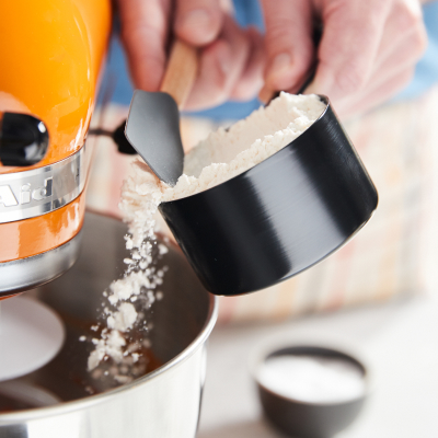 A person scraping flour into an orange KitchenAid® stand mixer bowl