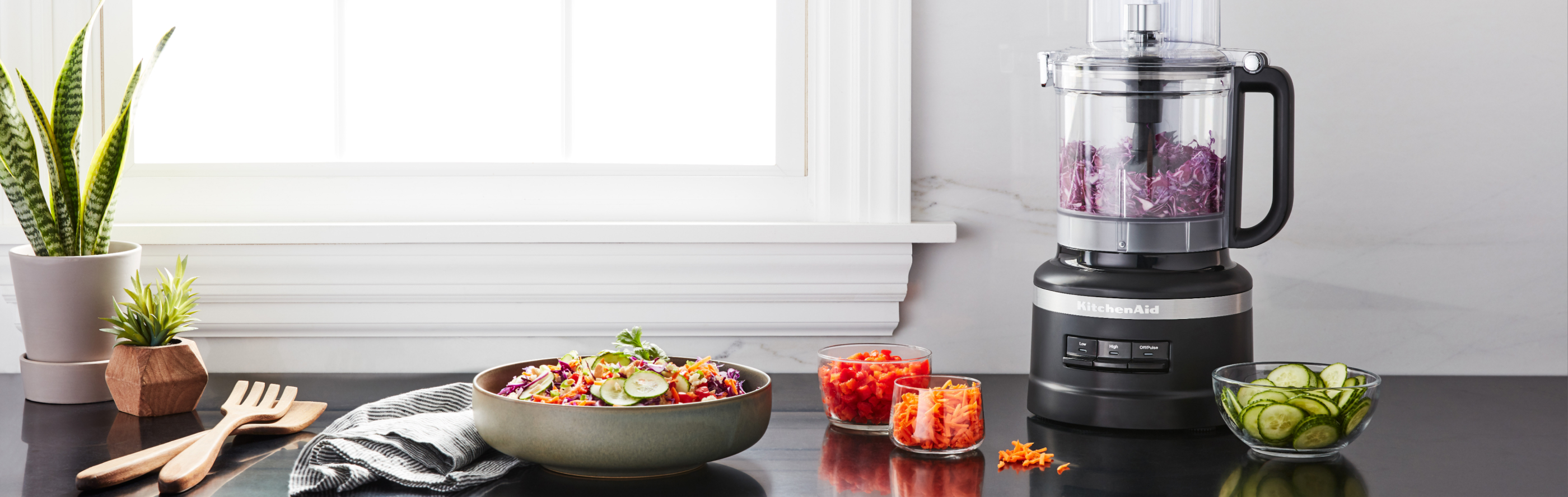 Black KitchenAid® food processor full of shredded red onion next to prepared veggies on the countertop