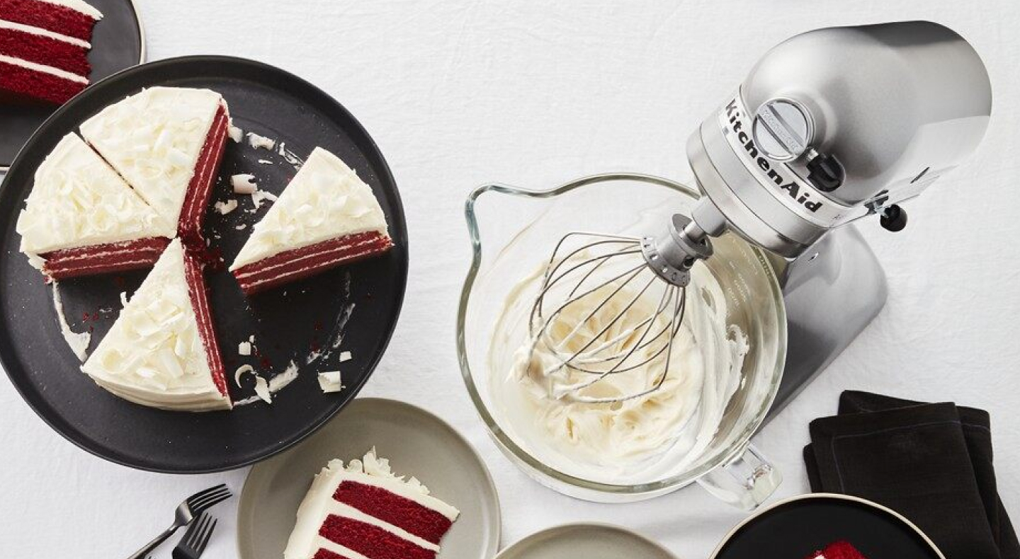 A KitchenAid® stand mixer with whipped white frosting next to a sliced red velvet cake.