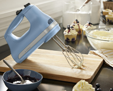 A KitchenAid 5-Speed Hand Mixer on a cutting board surrounded by frosting, jam and cupcakes.