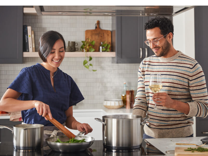 TitTwo people cooking together using several stainless steel pans on an induction cooktop.le Needed Two people cooking together using several stainless steel pans on an induction cooktop.