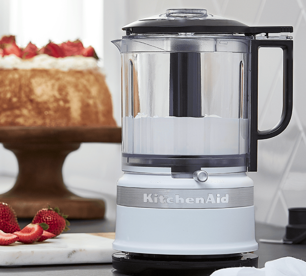 A KitchenAid® food processor in front of a cake topped with strawberries