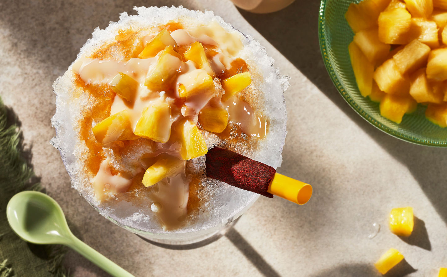 Shaved ice dessert topped with syrup and chopped fruit sitting next to green spoon Shaved ice dessert topped with syrup and chopped fruit sitting next to green spoon