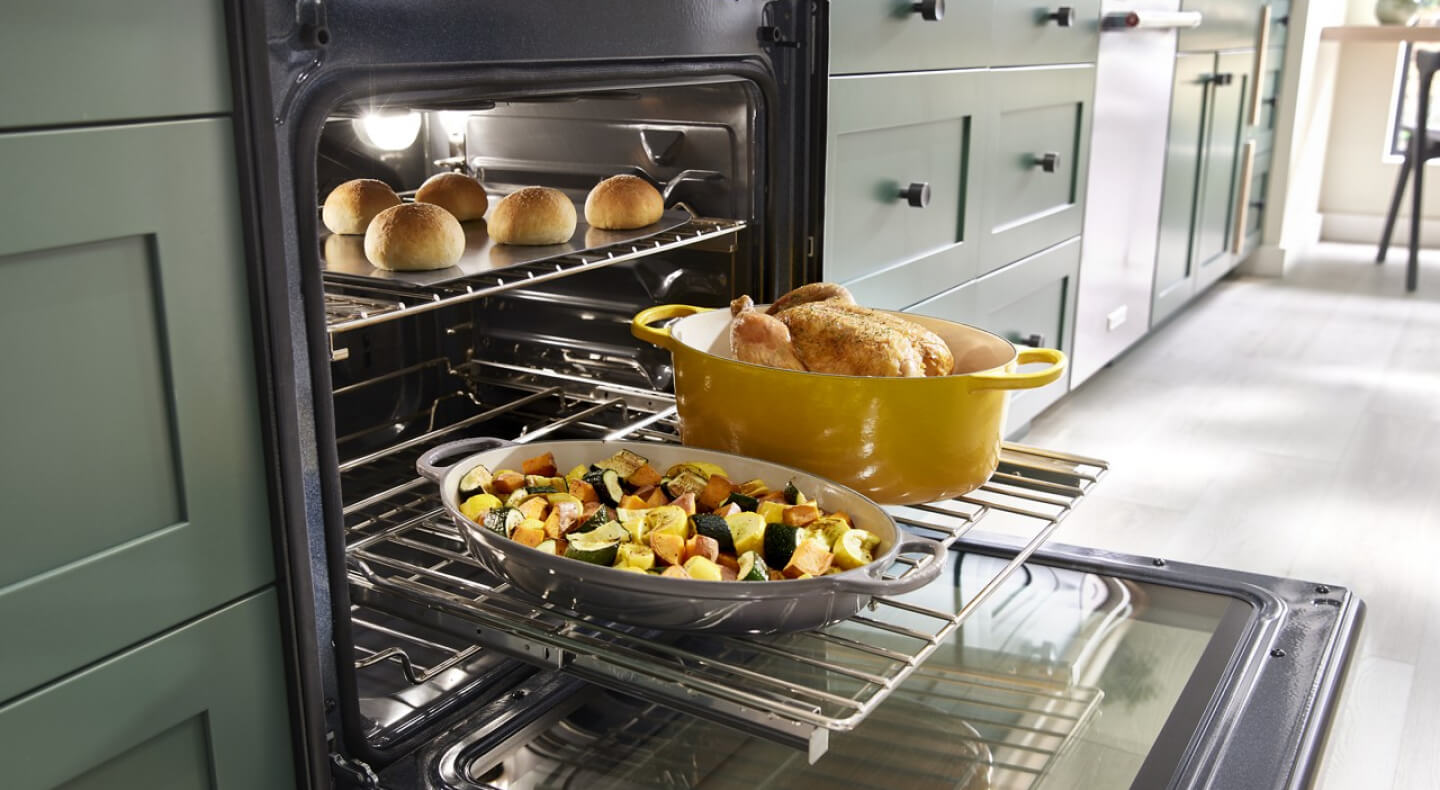 A close up of an open oven in a kitchen with a chicken, vegetables and bread rolls all cooking inside