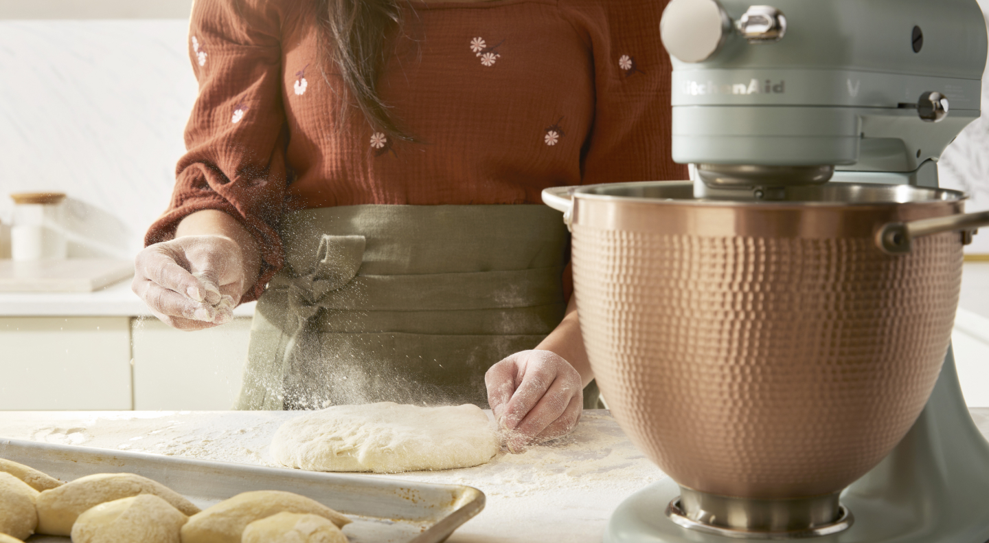 A person sprinkling dough with flour next to a KitchenAid® stand mixer. A person sprinkling dough with flour next to a KitchenAid® stand mixer.