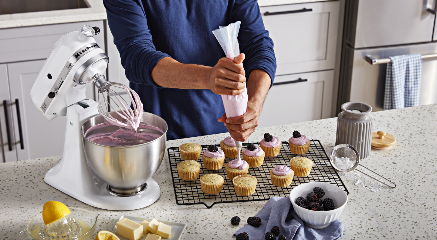 Person piping frosting onto cupcakes next to white stand mixer full of frosting Person piping frosting onto cupcakes next to white stand mixer full of frosting