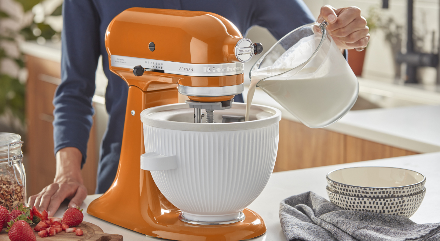 Person pouring milk into the white bowl of an orange stand mixer Person pouring milk into the white bowl of an orange stand mixer
