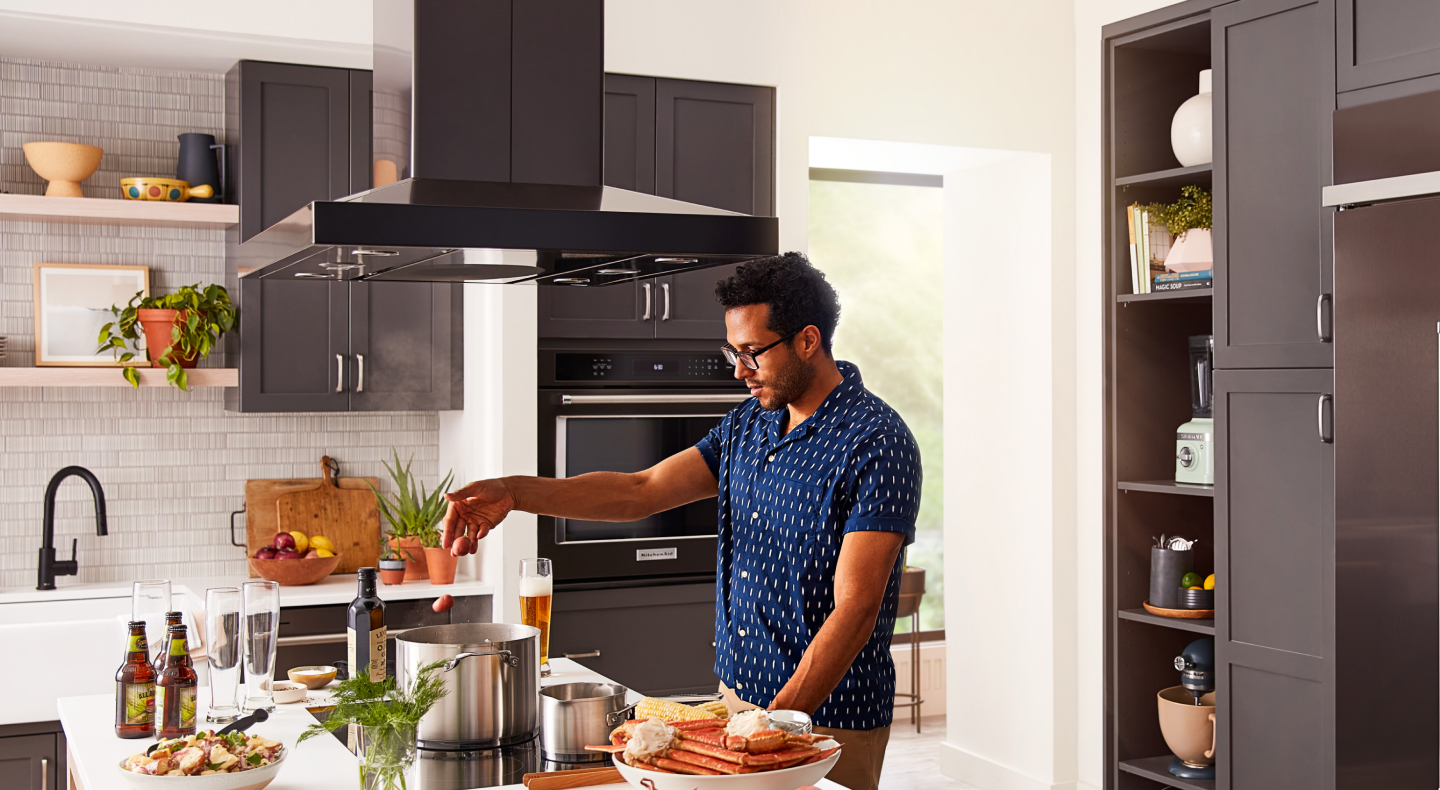 A person adding food to a pot on a KitchenAid® cooktop with a range hood above A person adding food to a pot on a KitchenAid® cooktop with a range hood above