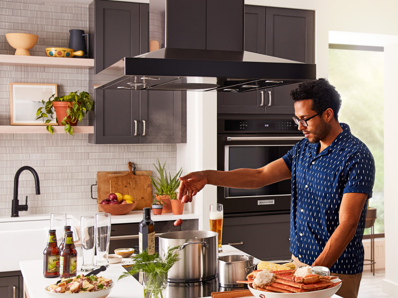 A person adding food to a pot on a KitchenAid® cooktop with a range hood above A person adding food to a pot on a KitchenAid® cooktop with a range hood above