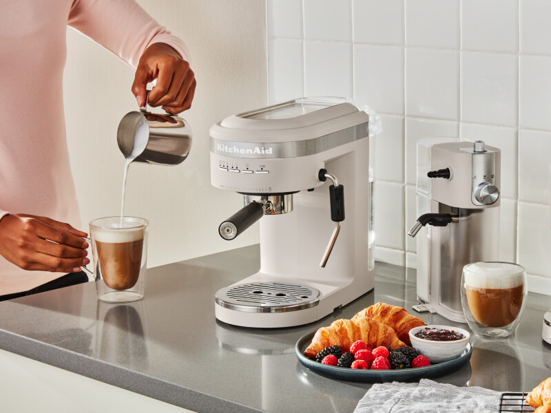 Person pouring frothed milk into an espresso drink next to an semi-automatic type of espresso machine Person pouring frothed milk into an espresso drink next to an semi-automatic type of espresso machine