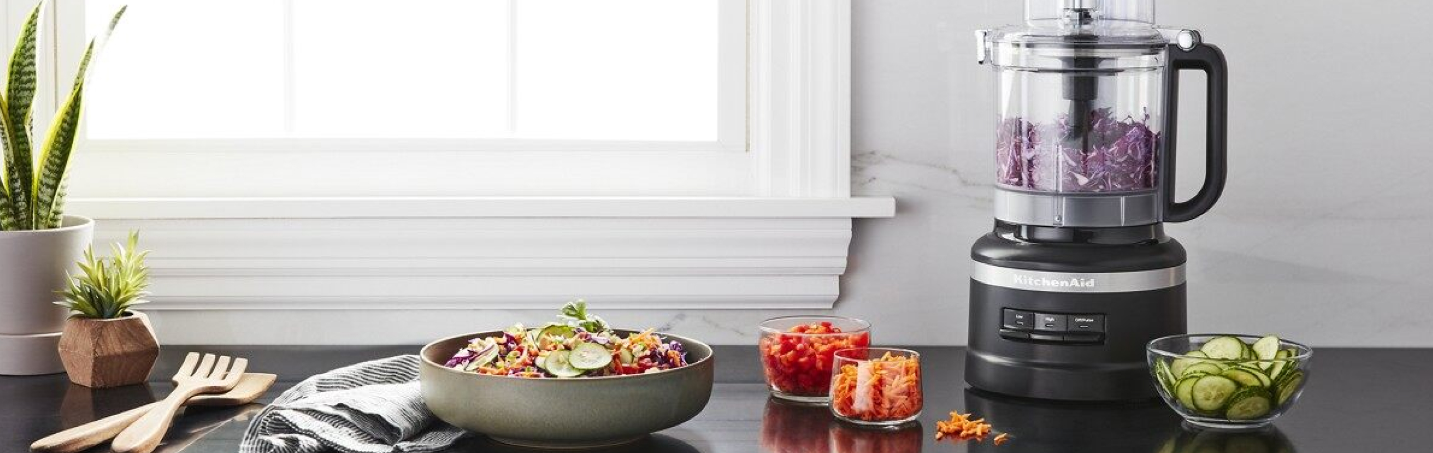 Black food processor on counter surrounded by processed vegetables and a bowl of salad