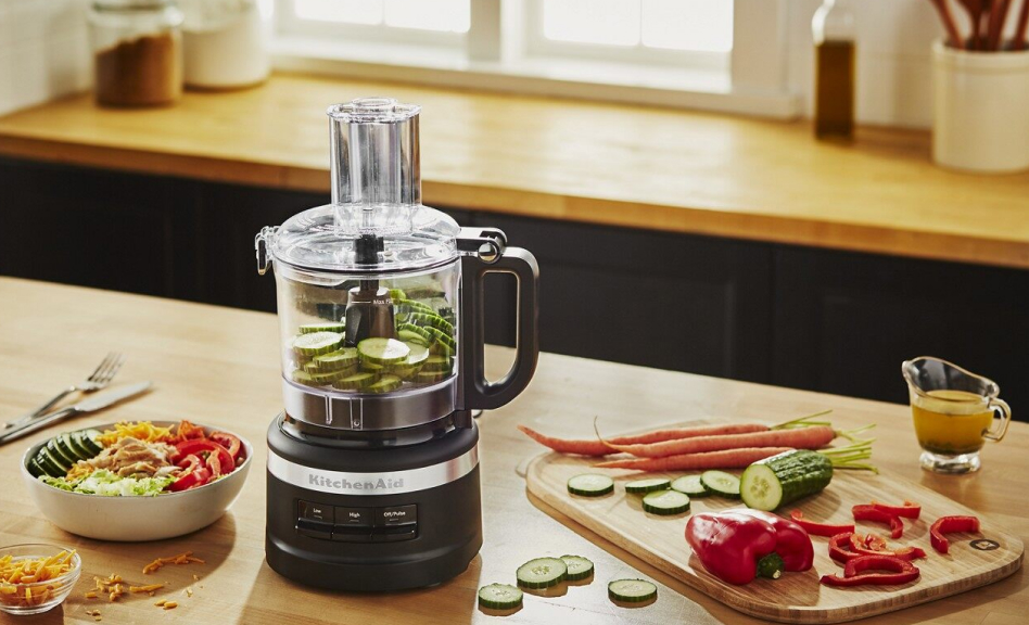 Black food processor with sliced cucumbers surrounded by vegetables and a salad Black food processor with sliced cucumbers surrounded by vegetables and a salad