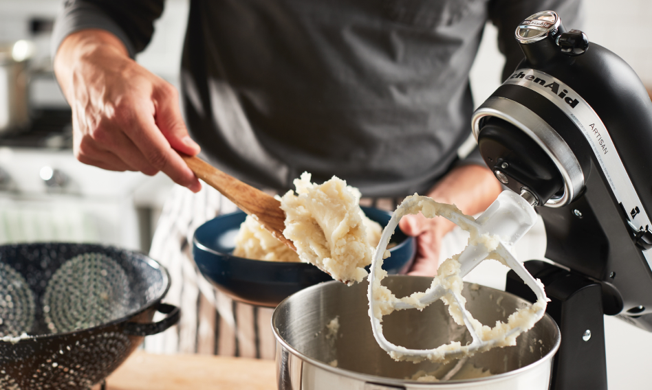 Male hand holding a bowl and a wooden spoon filled with mashed potatoes over a KitchenAid brand stand mixer Male hand holding a bowl and a wooden spoon filled with mashed potatoes over a KitchenAid brand stand mixer