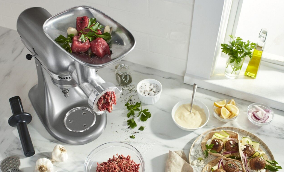 Silver KitchenAid brand stand mixer with uncooked beef inside food grinder attachment and condiments and spices on the side Silver KitchenAid brand stand mixer with uncooked beef inside food grinder attachment and condiments and spices on the side