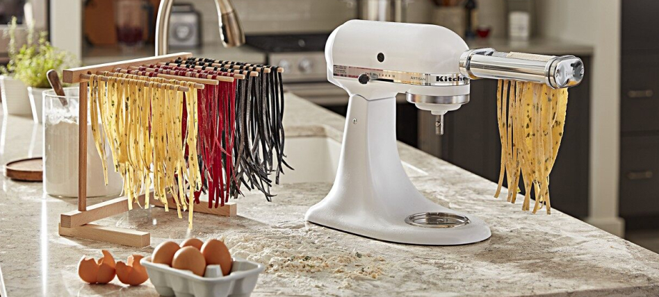 White KitchenAid brand stand mixer with pasta cutter attachment and wooden stand with uncooked pasta strips and eggs on a countertop White KitchenAid brand stand mixer with pasta cutter attachment and wooden stand with uncooked pasta strips and eggs on a countertop