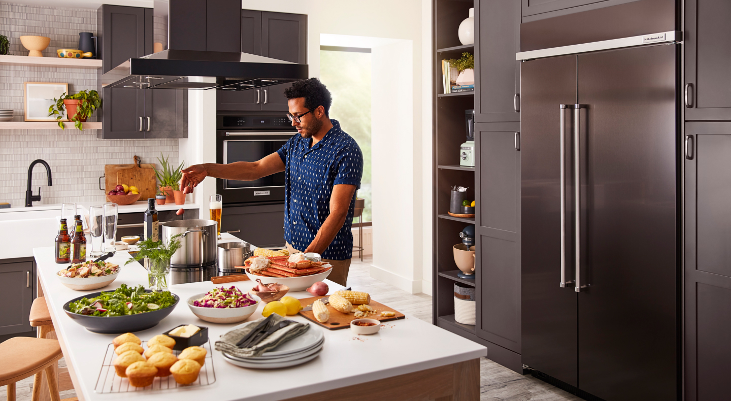 Person cooking at an island in front of a built-in, black stainless steel KitchenAid® side-by-side refrigerator Person cooking at an island in front of a built-in, black stainless steel KitchenAid® side-by-side refrigerator